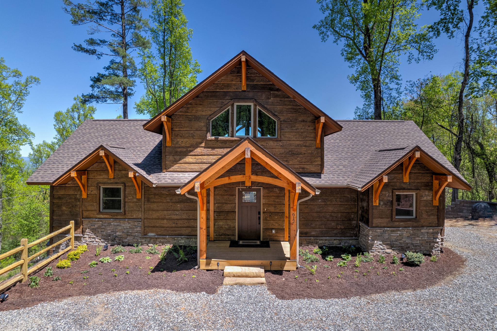 Your mountain arrival · A timber-framed entrance welcomes you to the cabin, surrounded by tall trees and a gravel arrival area.